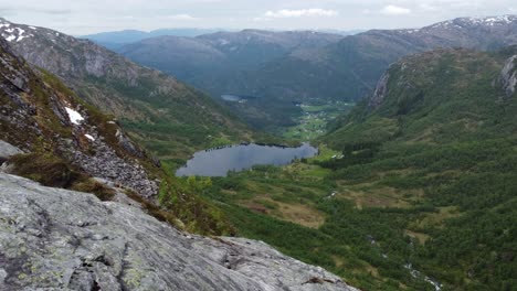 Leirovatnet-freshwater-lake-and-Eidslandet-village-in-background-is-revealed-from-a-distand-mountaintop-in-Stamnes-Vaksdal-Norway---Forward-moving-aerial-close-to-cliff-and-mountain-viewpoint