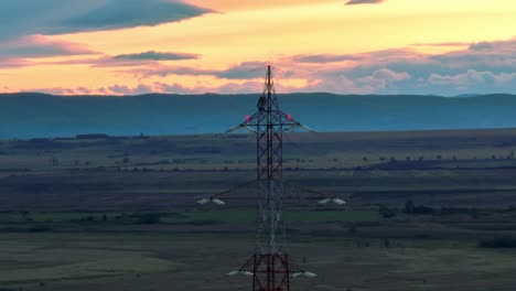 Aerial-view-showing-electricity-pylon-on-rural-field-in-front-of-hills-at-sunset