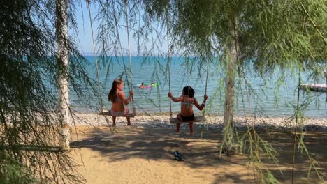 Back-view-of-two-little-girls-on-vacation-having-fun-swinging-on-seafront-rope-swing-with-tree-fronds-in-foreground