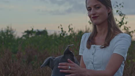 Portrait-of-attractive-young-woman-petting-the-head-of-her-pitbull-in-a-heather-field-at-dusk-and-looking-towards-camera