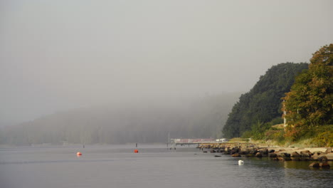 Peaceful-Morning-at-a-lake-with-fog-on-the-water-and-green-trees-on-the-shore-in-Scandinavia-at-fjord