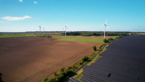 Aerial-trucking-shot-of-agriculture-field-with-installed-photovoltaic-units-and-spinning-wind-turbines-during-clear-sky-in-sun