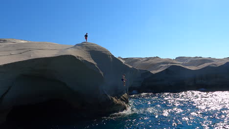 Two-people-stand-on-the-highest-point-of-a-cliff,-one-jumps-into-the-water-of-the-Mediterranean-sea
