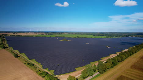 Aerial-backwards-shot-of-modern-solar-farm-field-installed-in-rural-field-during-sunlight-and-blue-sky---4k-footage