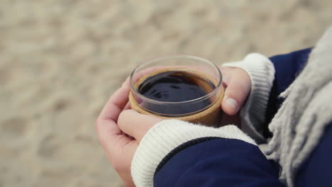 Woman-holding-hot-coffee-mug-in-her-hands-at-a-beach-on-a-cold-day-to-warm-herself