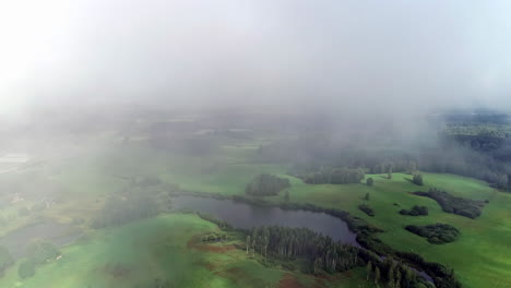 Low-valley-fog-over-a-lush,-green-landscape-with-ponds-and-copse-of-trees---aerial-flyover
