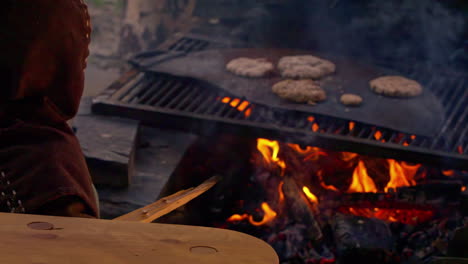 Barbecue-in-Medieval-Era,-Man-in-Authentic-Clothes-Controlling-the-Fire-Under-Meat-on-Metal-Plate,-Slow-Motion