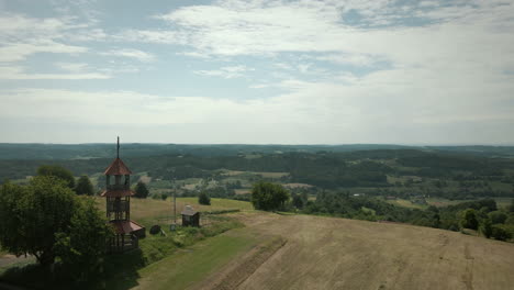 Panorama-Landschaftsaufnahme-Eines-Hügels-Mit-Kleinem-Turm-Auf-Der-Spitze-Und-Wald-Mit-Bäumen-Und-Grünem-Gras-Landwirtschaftliche-Felder-Um-Sie-Herum-Blauer-Himmel-Und-Hügel-Im-Hintergrund-In-Slowenien,-Drohnenaufnahme-Aus-Der-Luft