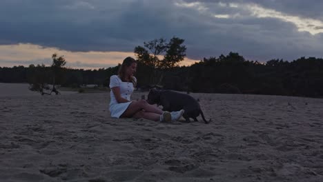 American-Staffordshire-Terrier-lying-down-next-to-young-woman-sitting-in-sand-dunes-at-dusk