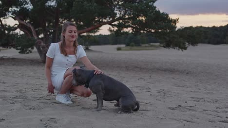 Junge-Frau,-Die-Ihren-Amerikanischen-Staffordshire-Terrier-Hund-In-Sanddünen-Streichelt-Und-In-Richtung-Kamera-Lächelt