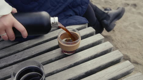 Woman-Pouring-Hot-Coffee-From-Thermos-Into-Mug-On-Wooden-bank-at-the-beach-In-Morning-in-slow-motion