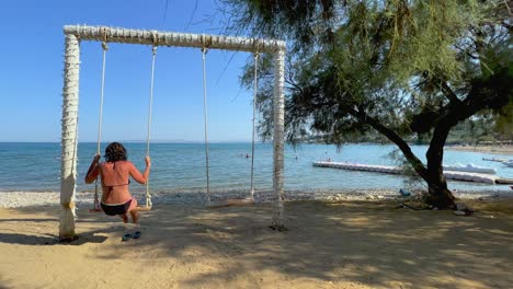 Back-view-of-adult-woman-on-summer-holiday-enjoy-swinging-on-rope-swing-looking-at-calm-sea