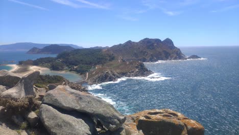 Mountainous-island-with-cliffs-and-beaches-and-camping-in-the-background,-sunny-day,-panoramic-shot-turning-right,-Cíes-Islands,-Pontevedra,-Galicia,-Spain