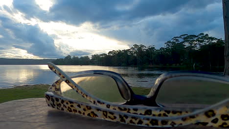 Reading-glasses-are-placed-on-a-picnic-table-overlooking-a-lake-at-sunset-on-a-cloudy-evening