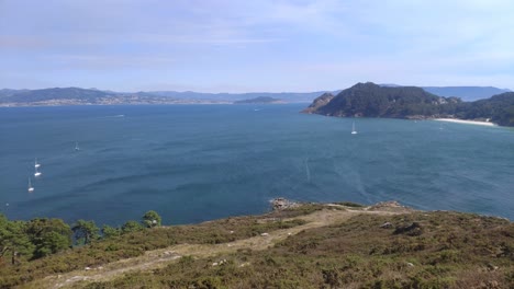 Elevated-view-of-the-sea-and-mountainous-island-with-beach-and-boats-sailing,-sunny-day,-panoramic-overhead-shot-turning-right,-Cíes-Islands,-Pontevedra,-Galicia,-Spain