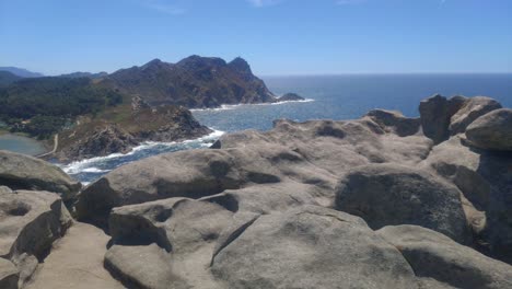 Close-up-of-eroded-granite-rock,-mountainous-island-with-cliffs-and-beaches-with-heat-mist,-sunny-day,-panoramic-shot-turning-left,-Cíes-Islands,-Pontevedra,-Galicia,-Spain