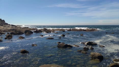 Low-sea-tide-with-reflections-of-the-sun-and-waves-crashing-against-the-cliff-rocks-on-a-cloudless-sunny-day,-shooting-traveling-to-the-rolling-left,-Cíes-Islands,-Pontevedra,-Galicia,-Spain