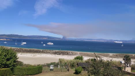 Fire-on-the-horizon-behind-the-coast-with-orchards-and-beach-with-luxury-yachts-and-boats-anchored-on-a-sunny-summer-day,-panoramic-shot-blocked,-Cíes-Islands,-Pontevedra,-Galicia,-Spain