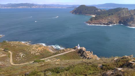 Elevated-view-of-the-road-to-the-lighthouse,-the-sea-and-the-mountainous-island-with-beach-and-boats-sailing,-sunny-day,-panoramic-shot-turning-right,-Cíes-Islands,-Pontevedra,-Galicia,-Spain