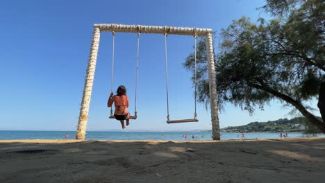 Low-angle-back-view-of-adult-woman-on-summer-holiday-enjoy-swinging-on-rope-swing-looking-at-calm-sea