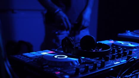 Kopfhörer-Werden-Auf-Ein-Digitales-DJ-Mischpult-Im-Innenbereich-Unter-Greller-Blauer-Beleuchtung-Gelegt,-Gefilmt-Als-Stationäre-Nahaufnahme-Mit-Sich-Bewegendem-DJ-Im-Hintergrund