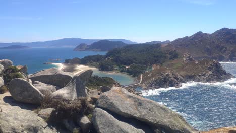 Mountainous-island-with-cliffs-and-beaches-and-camping-in-the-background,-sunny-day,-panoramic-high-shot-turning-right,-Cíes-Islands,-Pontevedra,-Galicia,-Spain