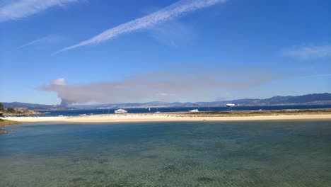 Fire-on-the-horizon-behind-the-coast-and-beach-with-luxury-yachts-and-boats-anchored-on-a-sunny-summer-day,-panoramic-shot-turning-right,-Cíes-Islands,-Pontevedra,-Galicia,-Spain