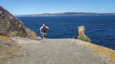 Man-without-clothes-with-backpack-comes-running-to-help-the-cliff-of-the-sea-with-the-coast-in-the-background-a-sunny-day-without-clouds,-Shot-blocked-low,-Cíes-Islands,-Pontevedra,-Spain