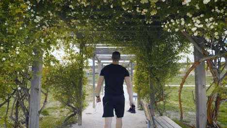 Panning-shot-of-an-athletic-man-walking-through-a-floral-archway-holding-gym-equipment
