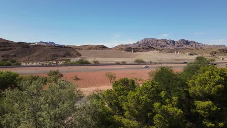 Vista-Lateral-De-La-Carretera-En-El-Desierto-Con-árboles-En-Primer-Plano,-Cielos-Azules-Y-Montañas-En-Segundo-Plano