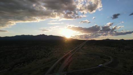 Aerial-View-Drone-Panning-Around-Highway-Exit-Ramp-with-Sun-Setting-Behind-Mountains-in-the-Background,-Light-Blue-Sky-and-Large-Clouds,-Lens-flare