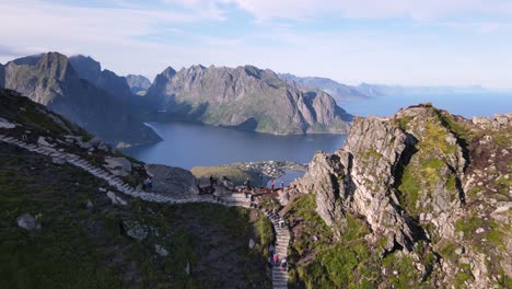 Mountain-hikers-walking-uphill-following-a-stony-staircase-trail-to-Reinebringen-peak,-people-enjoy-a-breathtaking-panorama-of-Reine-village-and-Lofoten-Islands-from-mount-ridge-viewpoint
