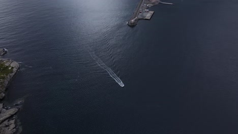 Top-tracking-view-of-Motorboat-entering-Reinevagen-bay-waters-passing-Reine-Lighthouse---orbiting-aerial-shot,-Norway