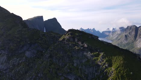 Aerial-flyover-passing-over-traveler-people-hiking-and-walking-on-Reinebringen-mountain-summit-ridge-revealing-numerous-steep-peaks-and-fjord,-Reine,-Norway