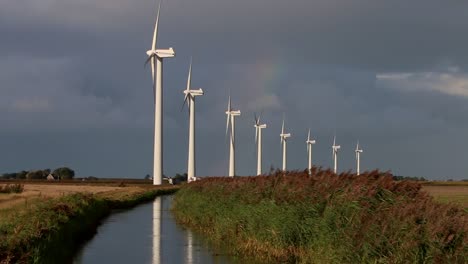 A-line-of-Wind-Turbines-turning-with-a-drainage-ditch-in-the-foreground-and-a-rainbow-and-stormy-sky