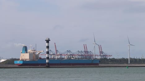 Commercial-ship-underway-with-dockyard-cranes,-and-wind-turbines-in-the-background