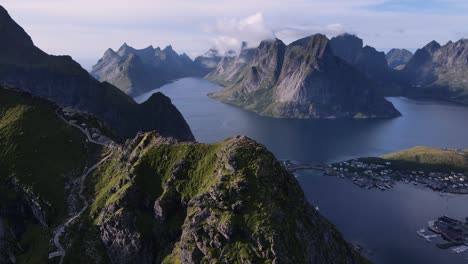 Reinebringen-mountain-ridge-summit-with-trail-walkways---aerial-orbiting-motion,-distant-view-of-Reine-village-at-the-mount-base-and-Lofoten-Islands-surrounded-by-still-water-lakes-and-fjords