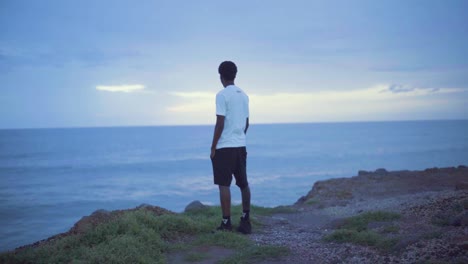 Young-black-man-standing-on-a-cliff-looking-at-the-sunset-and-the-ocean