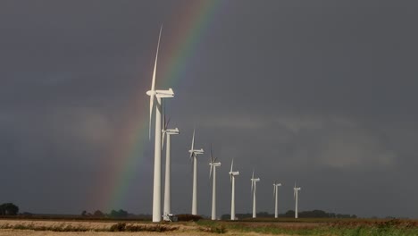 A-line-of-Wind-Turbines-turning-on-a-windy-day-with-a-stormy-sky-and-rainbow-in-the-background
