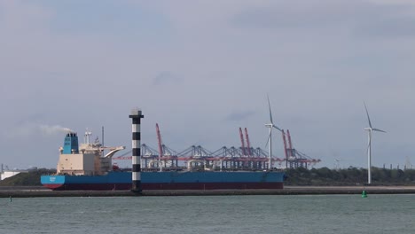 Commercial-ship-leaving-docks,-with-cranes-and-wind-turbines-in-the-background