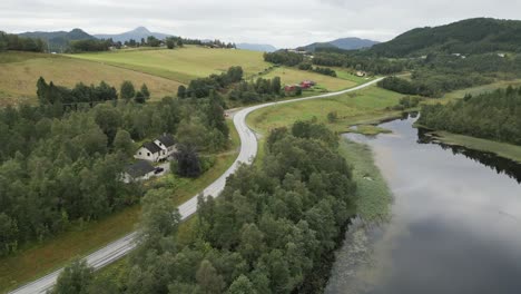 Movimiento-Ascendente-Desde-El-Tranquilo-Lago-Reflectante-En-Noruega-Sobre-La-Carretera-Hacia-Las-Verdes-Montañas-Cubiertas-De-Hierba-Con-Vistas-A-Las-Casas-Tradicionales-Rojas-Y-Amarillas