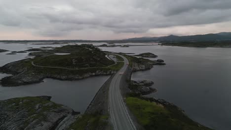 Flying-above-cars-passing-the-Atlantic-Ocean-Road-in-Norway-at-sunset-in-Summer-with-clouds-covering-the-mountains-in-the-distance-as-the-movement-goes-along-the-various-islands
