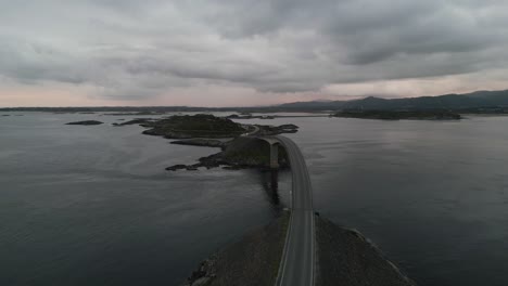 Slow-flying-movement-over-car-passing-the-Atlantic-Ocean-Road-in-Norway-at-sunset-with-the-headlights-turned-on-and-the-sunset-and-cloud-covered-mountains-in-the-background