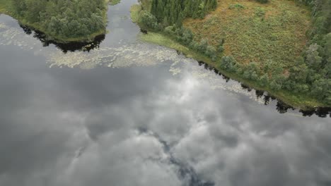 Downwards-tilted-aerial-view-of-clouds-reflecting-in-the-still-calm-lake-in-summer-Norway-with-green-grass-and-trees-being-revealed-at-the-end-of-the-scenery