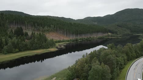 Flying-over-road-towards-the-extremely-calm-and-still-lake-showing-reflections-of-the-green-lush-trees-forest-in-Norway-with