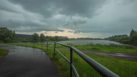 Cloudy-Sky-Over-Calm-Lake-And-Empty-Rural-Road-On-A-Rainy-Weather