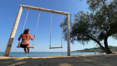 Low-angle-back-view-of-woman-on-summer-vacation-enjoying-relaxing-and-swinging-on-rope-swing-while-looking-at-calm-sea
