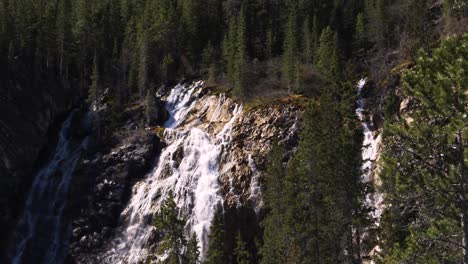 Wide-shot-of-cascading-waterfall-surrounded-by-pine-trees-on-sunny-mountainside