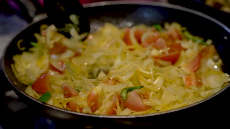 A-close-up-slow-motion-shot-of-a-frying-pan-as-a-chef-stirs-diced-onions,-chopped-tomatoes-and-chillies-around-a-hot-frying-pan-to-ensure-the-vegetables-are-cooked-evenly