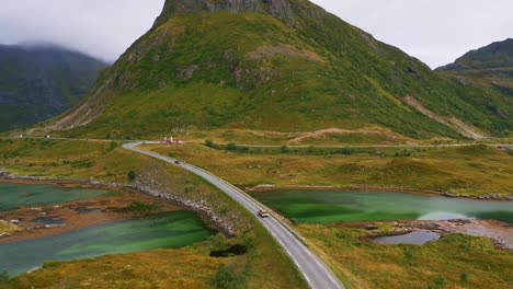 Following-a-car-driving-along-the-Fredvang-bridge-in-Lofoten-Northern-Norway-with-surrounding-yellow-green-grass-and-the-steep-mountain-in-the-background-with-red-little-huts-in-summer